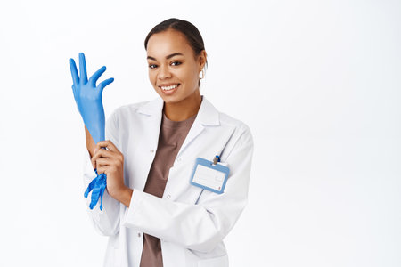 Doctor puts on gloves, prepare for patient checkup, smiling enthusiastic at camera, standing over white backgroundの写真素材