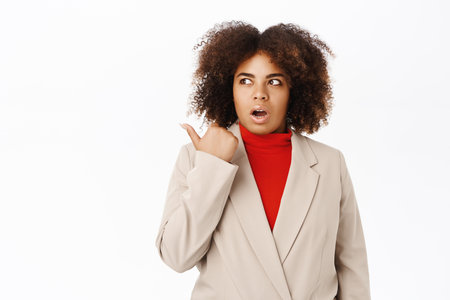 Shocked black businesswoman, pointing left, looking startled and speechless, standing in suit over white backgroundの写真素材