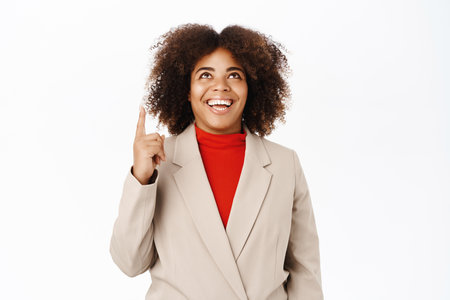 Smiling businesswoman, black woman, pointing finger up, shows direction on top, banner or company advertisement, stands over white backgroundの写真素材