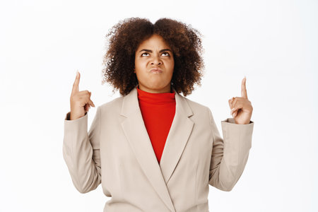 Disappointed, angry black saleswoman, pointing and looking up with jealous, displeased face expression, standing over white backgroundの写真素材