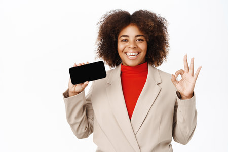 Smiling corporate woman shows horizontal mobile phone screen, company application, website, shows okay, stands over white backgroundの写真素材