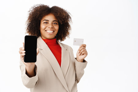 African american saleswoman, corporate woman shows her mobile phone screen and demonstrates credit card, stands over white backgroundの写真素材