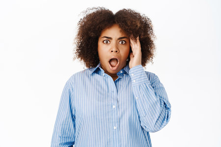 Portrait of black woman looking shocked and concerned at camera, gasps startled, stands over white studio backgroundの写真素材