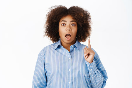 Eureka, she has an idea. Young african american woman has suggestion, lifting one finger up and looking excited, standing over white backgroundの写真素材