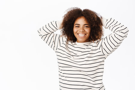 Carefree young african american woman in casual shirt, looking happy, smiling at camera, standing over white studio backgroundの写真素材