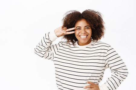 Smiling positive african ameircan girl shows peace, v-sign near eye and looks happy, stands over white backgroundの写真素材