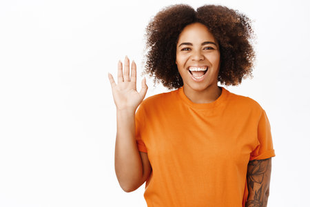 Friendly smiling african american woman saying hello, waving hand at you to greet, hi gesture, standing over white background in orange tshirtの写真素材