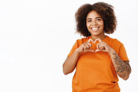 Portrait of smiling Black woman with curly hair, shows heart love sign, stands over white backgroundの写真素材