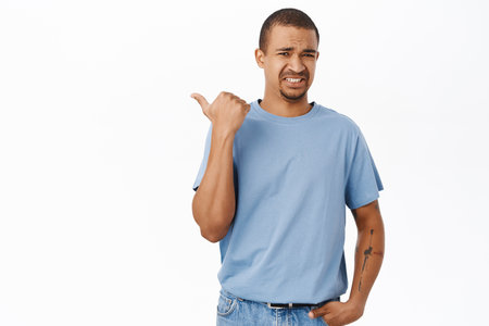 Displeased arab man points left, cringe and grimaces from dislike, stands over white backgroundの写真素材