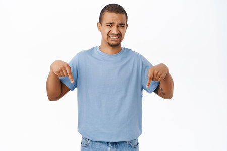 Disappointed man points fingers down with cringe grimace, dislike smth below, stands over white backgroundの写真素材