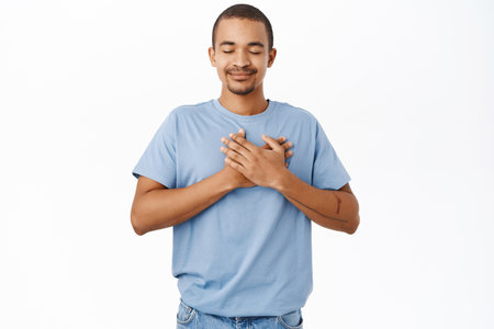 Smiling nice guy holds hands on heart, heartwarming and caring feeling, standing over white backgroundの写真素材