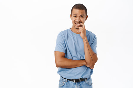 Nervous guy biting his finger and looking worried, having doubts, standing over white backgroundの写真素材