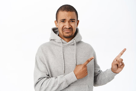 Crying upset guy pointing right, showing advertisement with disappointed face, standing in grey hoodie over white backgroundの写真素材