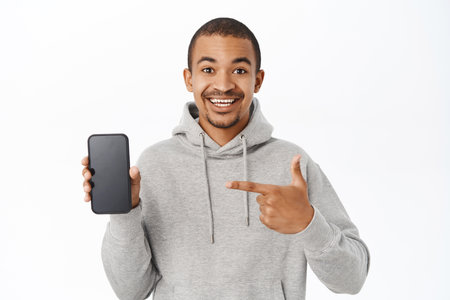 Handsome man shows mobile phone screen, points at telephone and smiles, recommends application, stands over white backgroundの写真素材