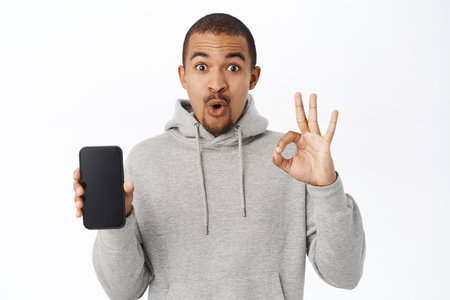 Amazed guy shows okay sign and mobile screen, smartphone app interface, recommends online store or application on telephone, standing over white backgroundの写真素材
