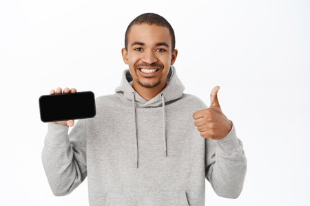 Enthusiastic arab guy shows mobile phone screen and thumbs up, recommends website, telephone application, stands over white backgroundの写真素材