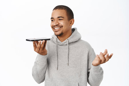 Handsome arab guy record voice message on smartphone, talking into mobile speakerphone, using telephone voice translation, standing over white backgroundの写真素材