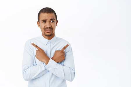Portrait of confused office worker, young man pointing sideways, cannot make decision, look clueless, standing over white backgroundの写真素材