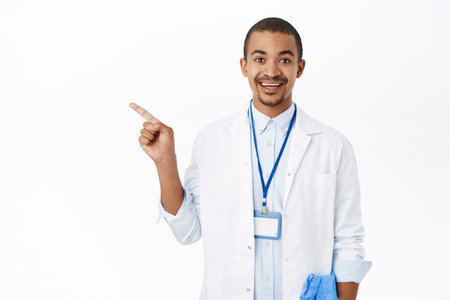 Portrait of young doctor pointing finger left, showing clinic logo, advertisement or price banner, standing over white backgroundの写真素材