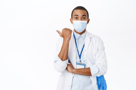 Surprised doctor in medical mask from covid, pointing left, looking amazed, standing over white backgroundの写真素材