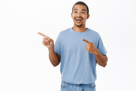 Portrait of smiling arab man pointing fingers left, shows direction, advertising, standing in blue tshirt over white backgroundの写真素材