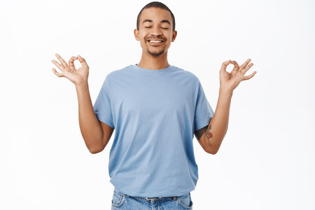 Mindfulness and inner peace. Smiling arab guy meditating, breathing air relaxed, resting, standing over white backgroundの写真素材