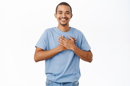 Smiling nice guy holds hands on heart, heartwarming and caring feeling, standing over white backgroundの写真素材