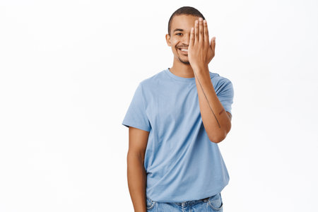 Enthusiastic young man hides one side of face, covers an eye and smiles, stands over white backgroundの写真素材