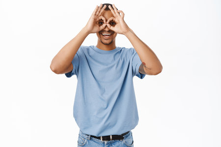Happy smiling guy looks through hand binoculars, okay signs on eyes, stands against white studio backgroundの写真素材