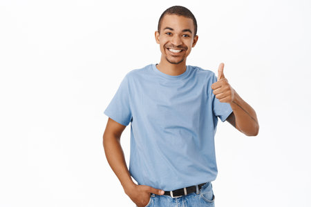 Cheerful young man shows thumbs up and smiling, approve, supports smth nice, stands over white backgroundの写真素材