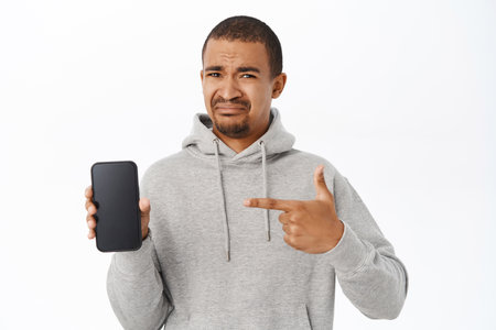 Disappointed guy points at his mobile phone screen, shows telephone and grimaces with dislike, looks upset, stands over white backgroundの写真素材