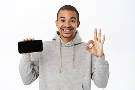 Amazed guy shows okay sign and mobile screen, smartphone app interface, recommends online store or application on telephone, standing over white backgroundの写真素材