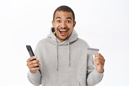 Excited man holding mobile phone and credit card, celebrating victory on smartphone, winning on telephone, standing over white backgroundの写真素材