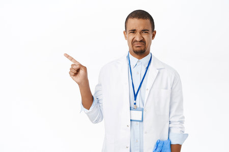 Sad male physician, doctor pointing left, grimacing and looking displeased, standing over white backgroundの写真素材