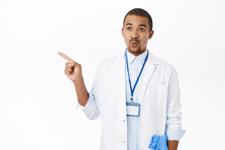 Doctor in white coat pointing finger left. Young intern in clinic showing way, demonstrating promo, standing over white backgroundの写真素材