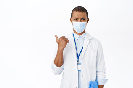 Shocked male physician, hospital doctor in medical mask looks concerned, points left at logo, copy space, stands over white backgroundの写真素材