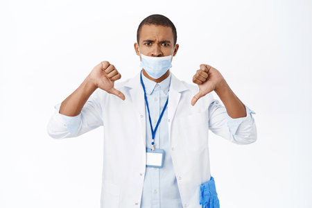 Doctor being upset with wrong way of wearing medical masks during covid-19 pandemic, shows thumbs down, stands over white backgroundの写真素材