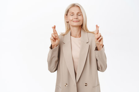 High hopes. Smiling blond mature woman, grandmother cross fingers and wishing, praying and hoping, standing over white backgroundの写真素材