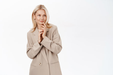 Beautiful mature woman thinking, looking with pleased smile and thoughtful face, wearing stylish french coat, standing over white backgroundの写真素材