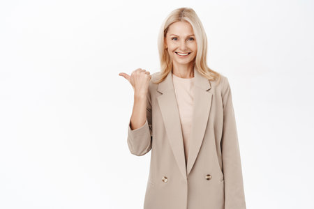 Portrait of beautiful blond mature woman, pointing finger left, showing copy space advertisement, standing over white backgroundの写真素材