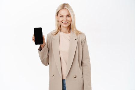 Online shopping. Smiling elegant blond lady, senior woman showing mobile phone screen app, smartphone application interface, standing over white backgroundの写真素材