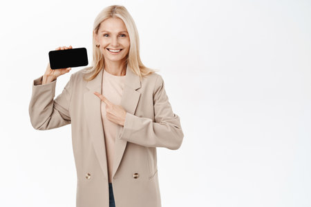 Portrait of blond elegant woman, pointing finger at horizontal smartphone screen, showing mobile phone and smiling, promoting website, online shopping, white backgroundの写真素材