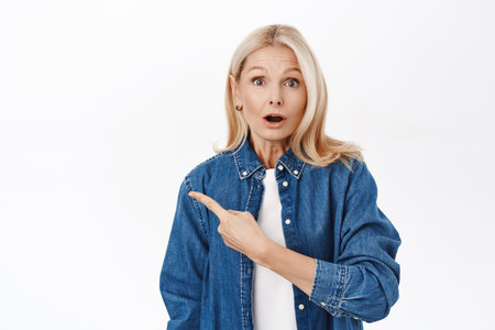 Close up portrait of surprised elegant senior woman, 50s years, pointing finger left, showing advertisement, promo offer, standing over white backgroundの写真素材