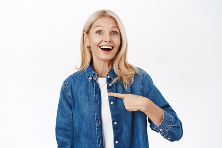 Portrait of excited smiling middle aged woman, senior lady pointing at herself with amazed face, standing over white backgroundの写真素材