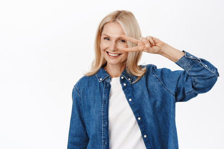 Portrait of smiling blond senior woman, 50 years, shows peace, v-sign gesture and looking happy, standing over white backgroundの写真素材