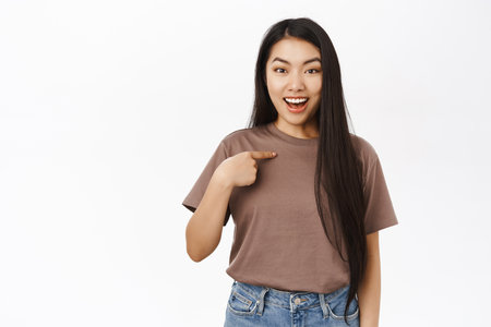 Beautiful smiling asian woman with long hair, points at herself and looks confident, self-promoting, standing over white backgroundの写真素材