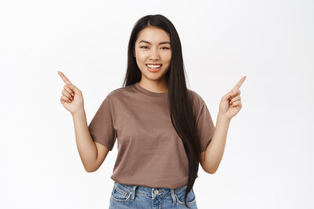 Portrait of beautiful asian female model pointing sideways, showing two ways, variants, stands over white backgroundの写真素材