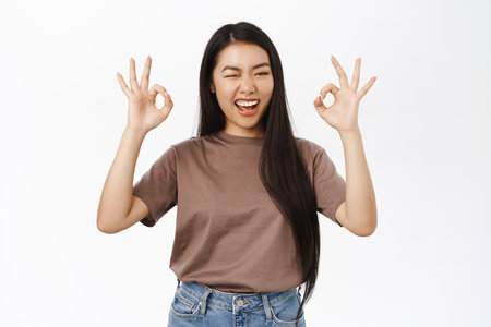 Portrait of enthusiastic chinese girl shows okay, ok sign in approval, winks at you, praise excellent choice, recommends, stands over white backgroundの写真素材