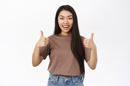 Smiling beautiful asian girl shows thumbs up, approve and like gesture, praise you, stands over white backgroundの写真素材