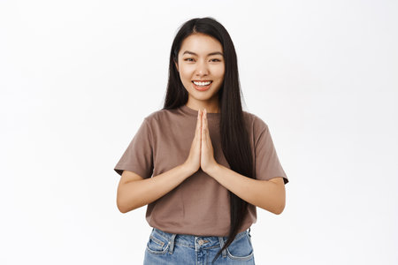 Smiling asian woman praying, holding hands in namaste, thank you gesture, standing relaxed, white studio backgroundの写真素材
